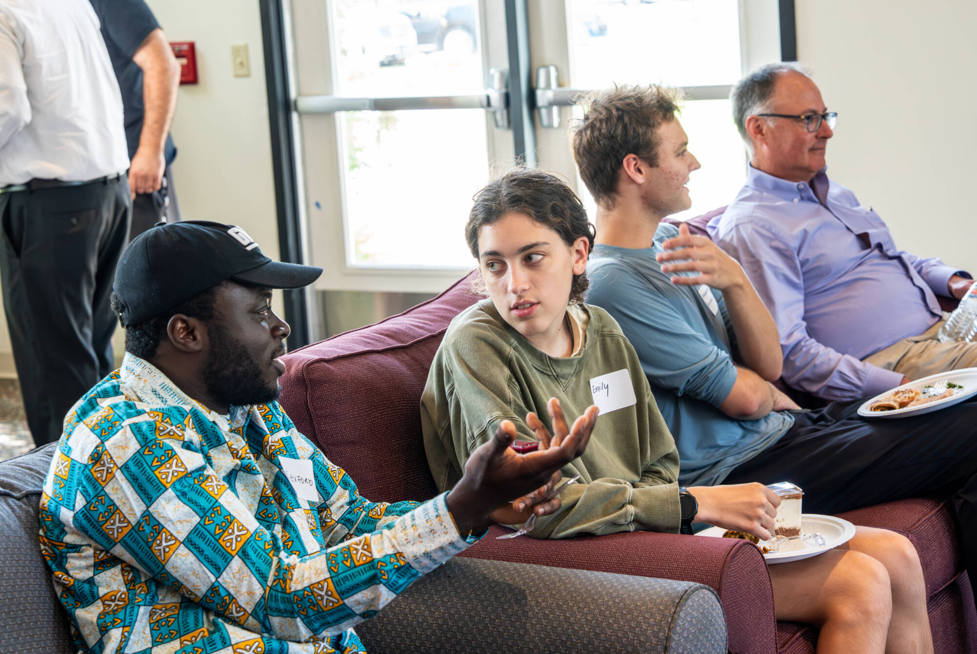 Students and faculty sit together on couches during a College of Computing mixer at GVSU, sharing conversation while enjoying food in a relaxed and welcoming setting.